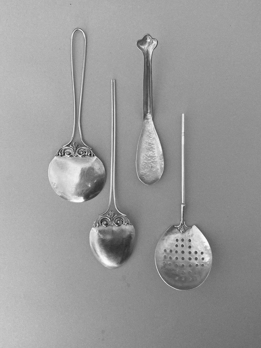 Four vintage metal utensils displayed on a flat surface, including two ornate tea strainers and two spoons, one perforated, in varying shapes and designs, with the label Tea Strainer.