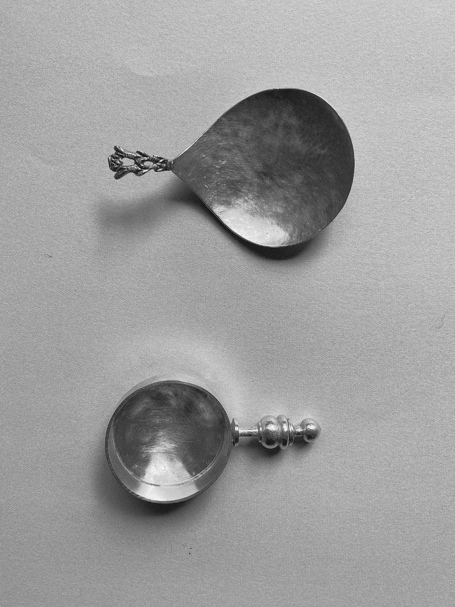 Two vintage metal Tea Spoons with ornate handles, one with a teardrop-shaped bowl and the other with a round bowl, placed on a light surface.