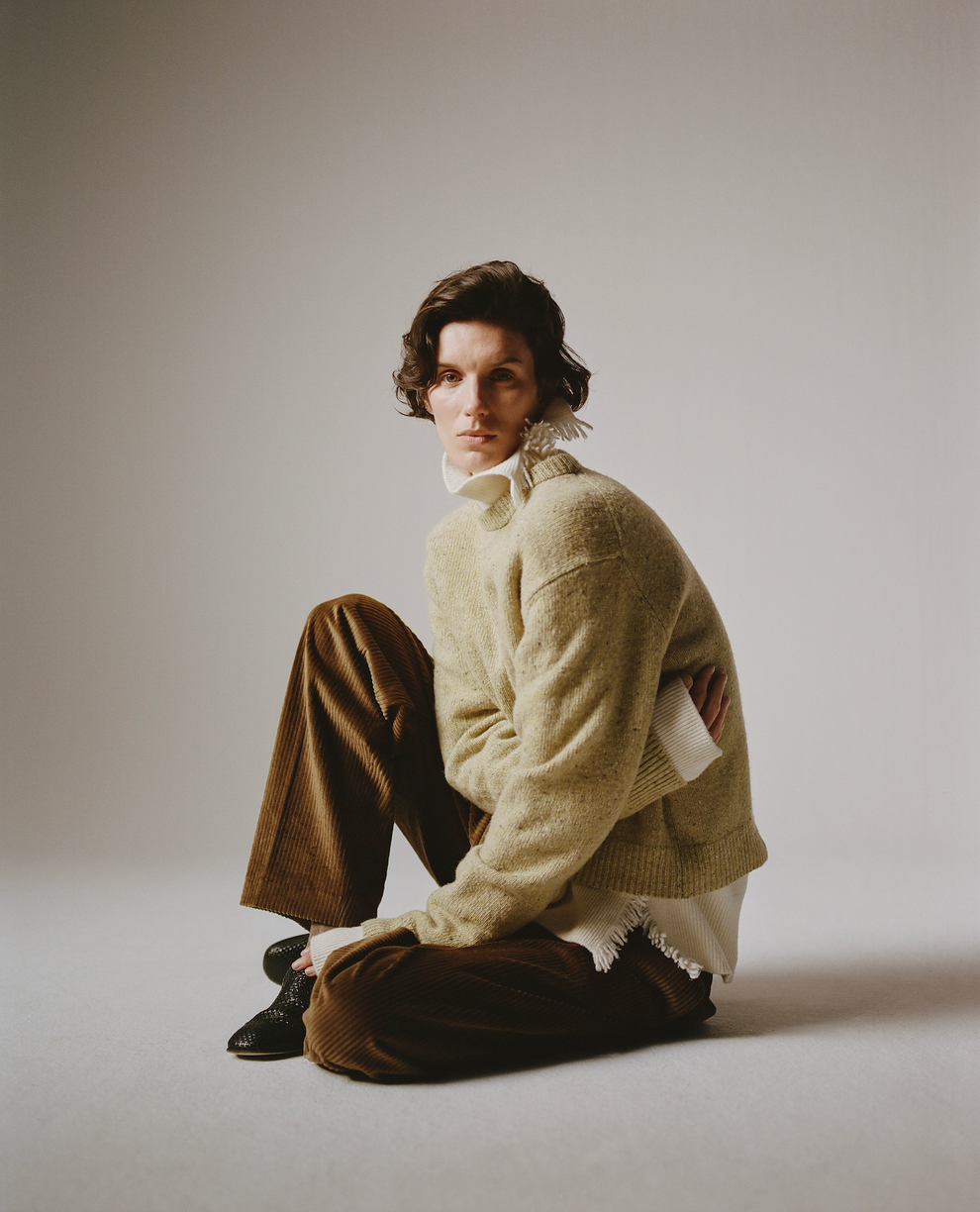 Person with short curly dark hair seated in a studio wearing a cream textured sweater over a white shirt brown trousers and black shoes against a neutral backdrop