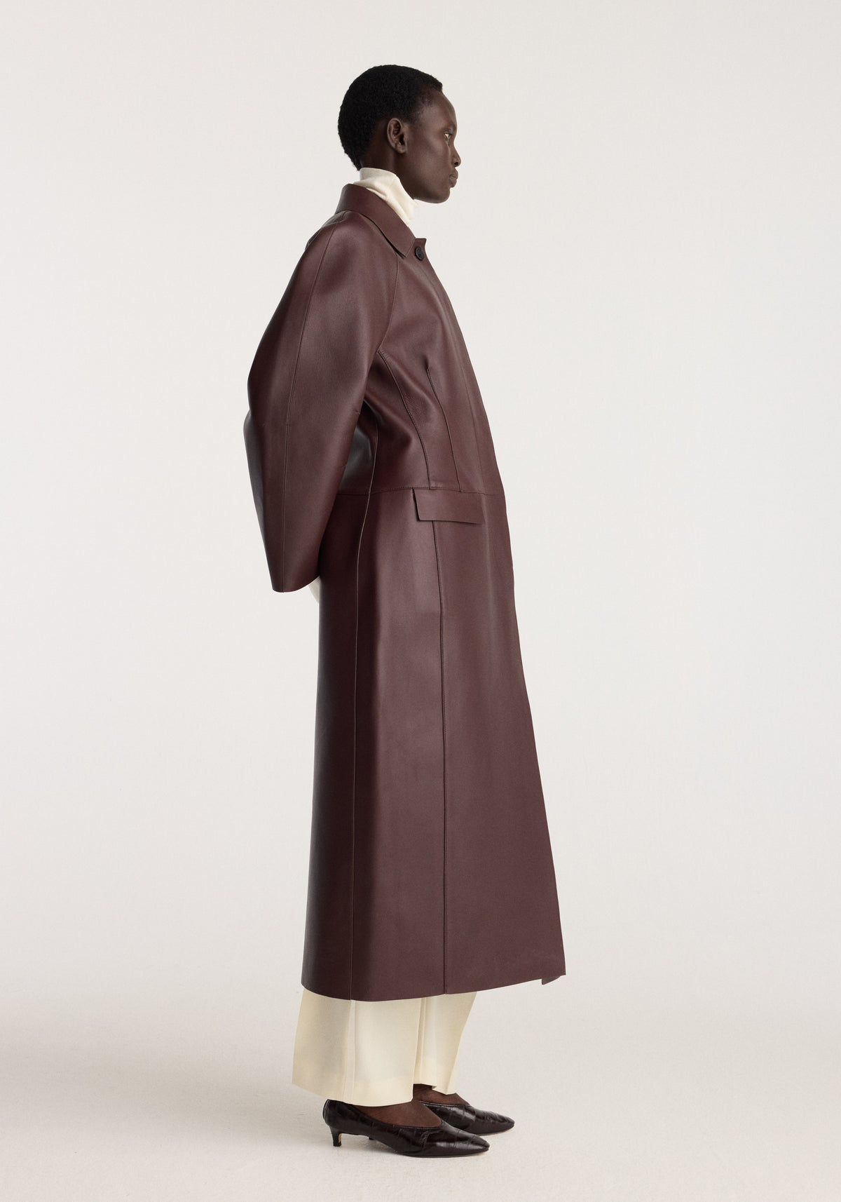 Sculptural Bonded Leather Coat | mahogany shown on a model in profile with a long mahogany leather silhouette and cream trousers over dark shoes