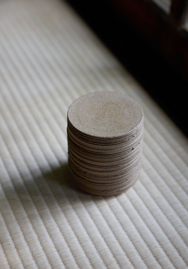 Stack of sand-colored discs on a woven mat, part of the Diplomatico Scented Candle set, featuring Róhe’s aromatic fragrance in a handmade ceramic vessel.
